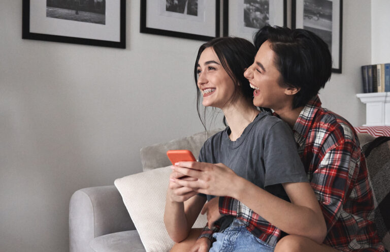 Young,Happy,Lesbian,Couple,Hugging,,Laughing,,Holding,Smartphone,At,Home.