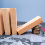 Wooden,Dominoes,Fall,On,A,Pile,Of,Coins,With,House