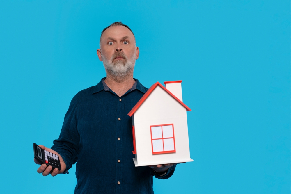 Shocked,Man,Holding,Model,House,And,Calculator,On,Blue,Blackground