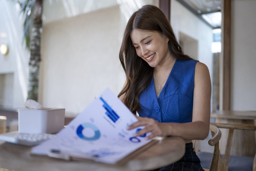 Young,Asian,Woman,Smiling,While,Reviewing,Business,Reports,And,Infographics