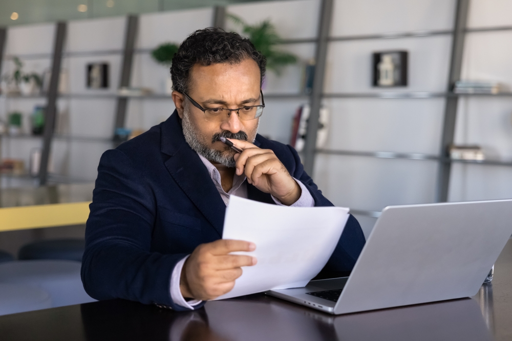 Thoughtful,Frowning,Senior,Indian,Businessman,Reading,Paper,Document,At,Laptop,