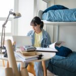 Student,Asian,Woman,Sitting,On,Bed,In,Dorm,Room,College