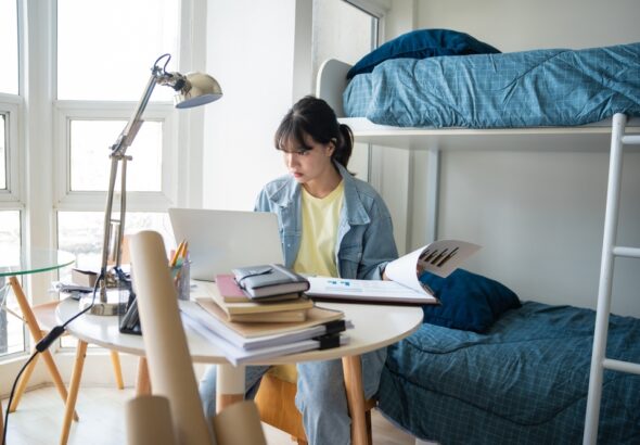 Student,Asian,Woman,Sitting,On,Bed,In,Dorm,Room,College
