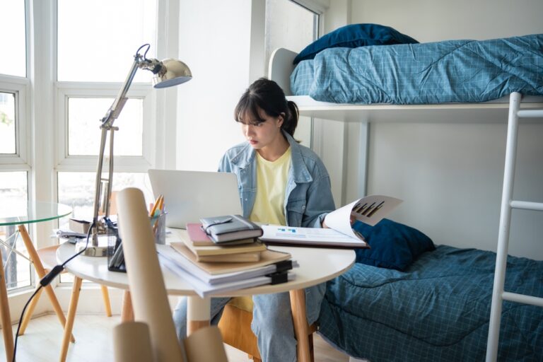 Student,Asian,Woman,Sitting,On,Bed,In,Dorm,Room,College