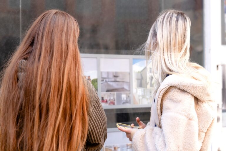 Two,Women,Stand,Close,Together,Looking,At,Art,Displays,On